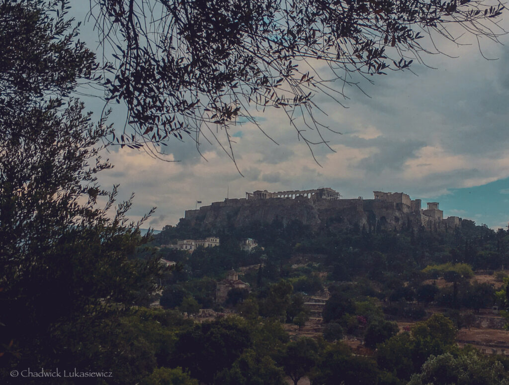 A distant view of the Acropolis of Athens framed by the branches of olive trees in the foreground. The ancient site, perched atop a rocky hill, features iconic structures such as the Parthenon and the Erechtheion, partially illuminated under a cloudy sky. The surrounding greenery and scattered historical ruins on the lower slopes add depth to the scene, emphasizing the blend of natural beauty and architectural grandeur.