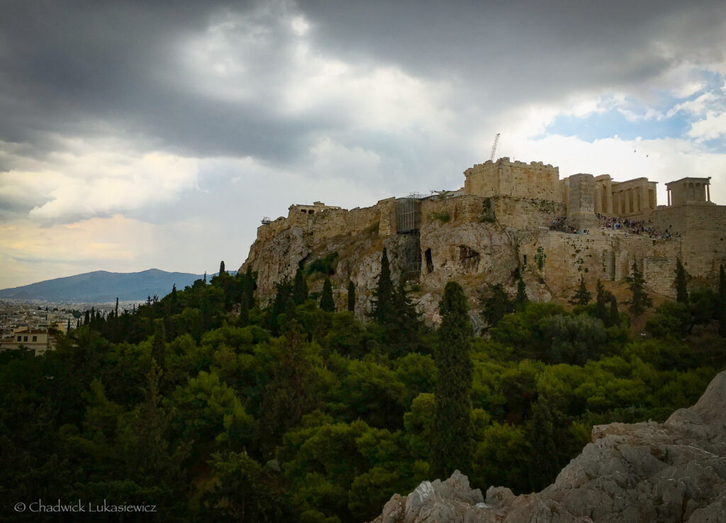 A majestic view of the Acropolis in Athens, Greece, perched atop a rocky hill surrounded by dense green trees. The ancient stone structures, including the Parthenon and the Erechtheion, are partially visible and undergoing restoration with scaffolding in some areas. The cloudy sky casts a dramatic atmosphere over the scene, with patches of sunlight breaking through to illuminate parts of the ruins and the lush hillside. The distant mountains and city skyline provide a stunning backdrop, emphasizing the historical and cultural significance of this iconic site.