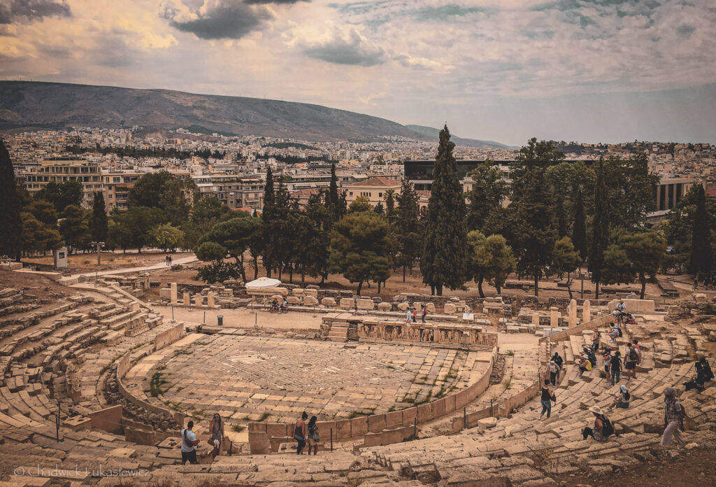 A panoramic view of the Theatre of Dionysus in Athens, Greece, located on the south slope of the Acropolis. The semi-circular stone seating, worn by centuries of use, surrounds a central stage area with remnants of ancient decorative carvings. Visitors are scattered across the tiers, exploring the historic site. Beyond the theater, lush trees and modern city buildings stretch out into the distance, with the rolling hills of Athens providing a scenic backdrop. The cloudy sky adds a dramatic atmosphere, blending the ancient and contemporary elements of the landscape.