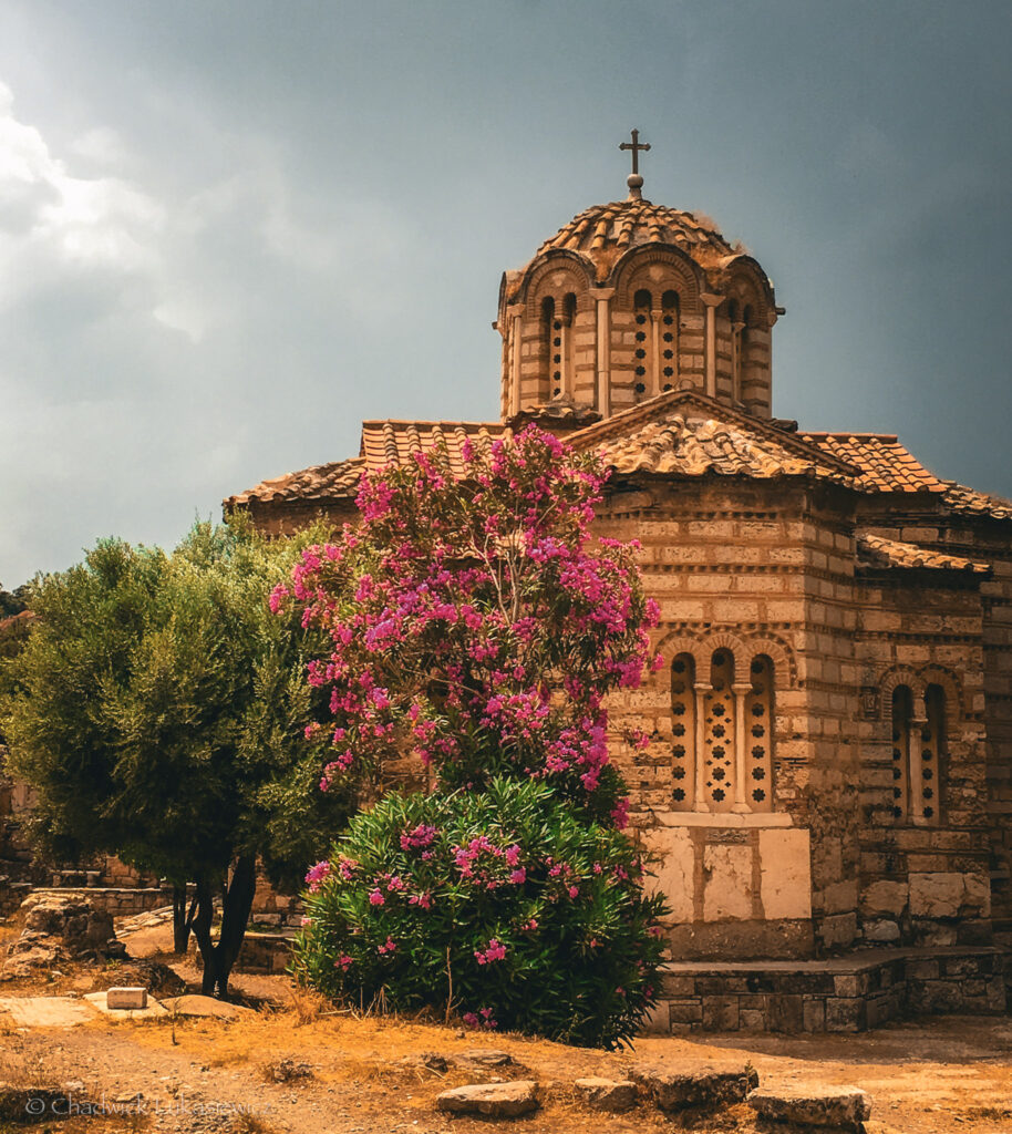 A picturesque view of the Church of the Holy Apostles in Athens, Greece, a small Byzantine-era church with a tiled dome topped by a cross. The church’s stone and brick facade is adorned with decorative arches and narrow windows. In the foreground, vibrant pink and purple flowers bloom on a lush bush, contrasting beautifully with the surrounding olive trees and the warm golden tones of the dry grass. The overcast sky adds a dramatic touch to the serene and historic setting, emphasizing the timeless charm of this ancient site.