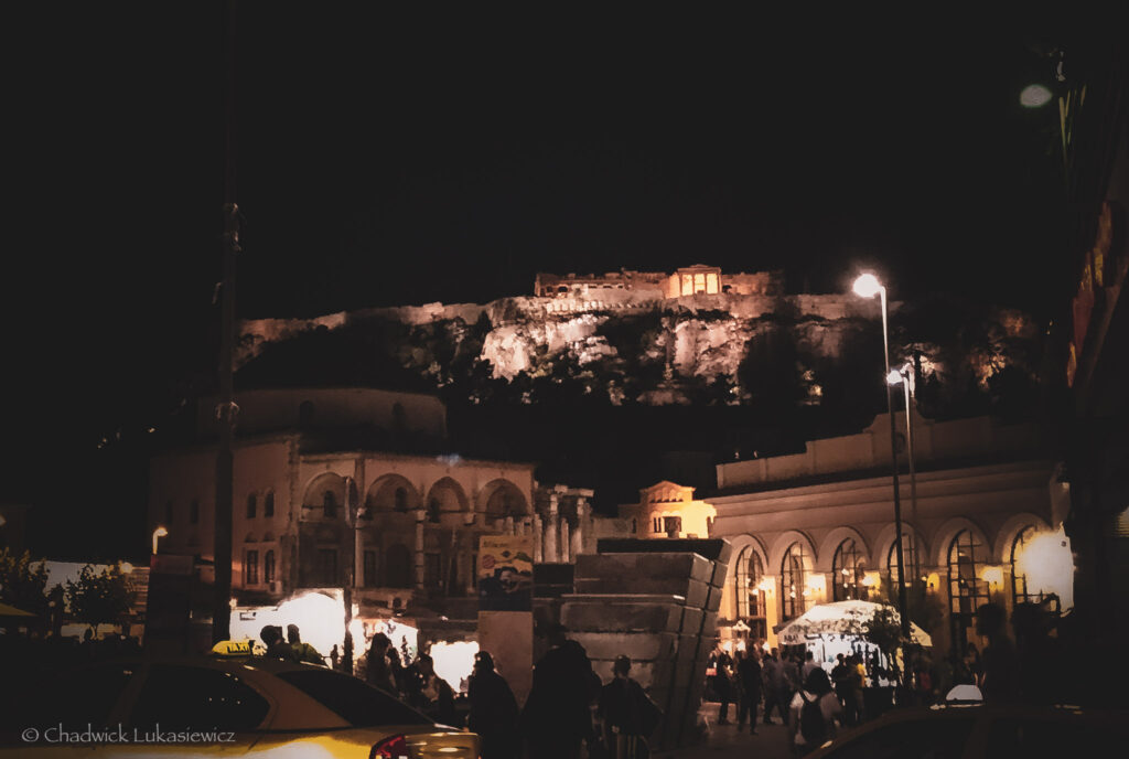 A nighttime view of Monastiraki Square in Athens, Greece, bustling with activity. In the foreground, a yellow taxi and silhouettes of people are visible, creating a lively urban atmosphere. To the left, the Tzistarakis Mosque, an Ottoman-era structure, stands illuminated by warm lights. The backdrop features the iconic Acropolis of Athens, dramatically lit against the dark sky, highlighting the ancient Parthenon atop the hill. The scene captures the intersection of modern life and historical heritage, emphasizing the vibrant energy of the square.