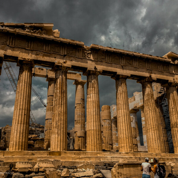 A dramatic view of the Parthenon in Athens, Greece, under a stormy sky, showcasing the iconic ancient columns and architectural details. The scene captures the awe-inspiring grandeur of the historic ruins, with two visitors in the foreground admiring the site. A crane in the background highlights ongoing preservation efforts, adding context to the timeless significance of this UNESCO World Heritage site. Ideal for an ADHD travel guide to Rome and Athens, emphasizing rich history and breathtaking landmarks