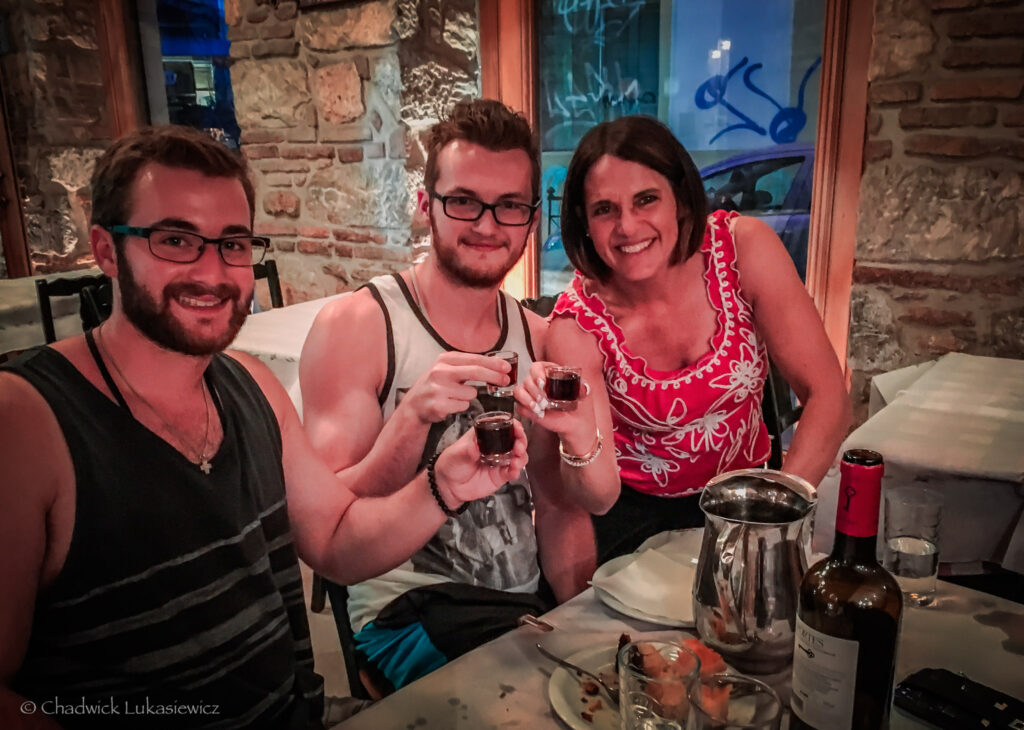 Three smiling people seated at a table in a cozy restaurant with stone walls, enjoying a meal together in Athens, Greece. Each person holds a small glass of tsipouro, a traditional Greek distilled spirit, raised for a toast. The table is set with a partially empty bottle of wine, a stainless steel carafe, glasses of water, and plates with remnants of food. The warm and intimate ambiance of the restaurant is enhanced by the soft lighting and visible details of the rustic decor.