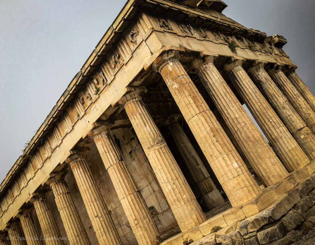 A close-up, angled view of the Temple of Hephaestus in Athens, Greece. The well-preserved Doric columns, made of weathered stone, showcase intricate fluting and support a detailed frieze adorned with carvings of mythological figures and motifs. The light golden hue of the ancient marble contrasts with the muted gray sky, highlighting the temple’s architectural beauty. This perspective emphasizes the temple’s imposing structure and its enduring historical significance.