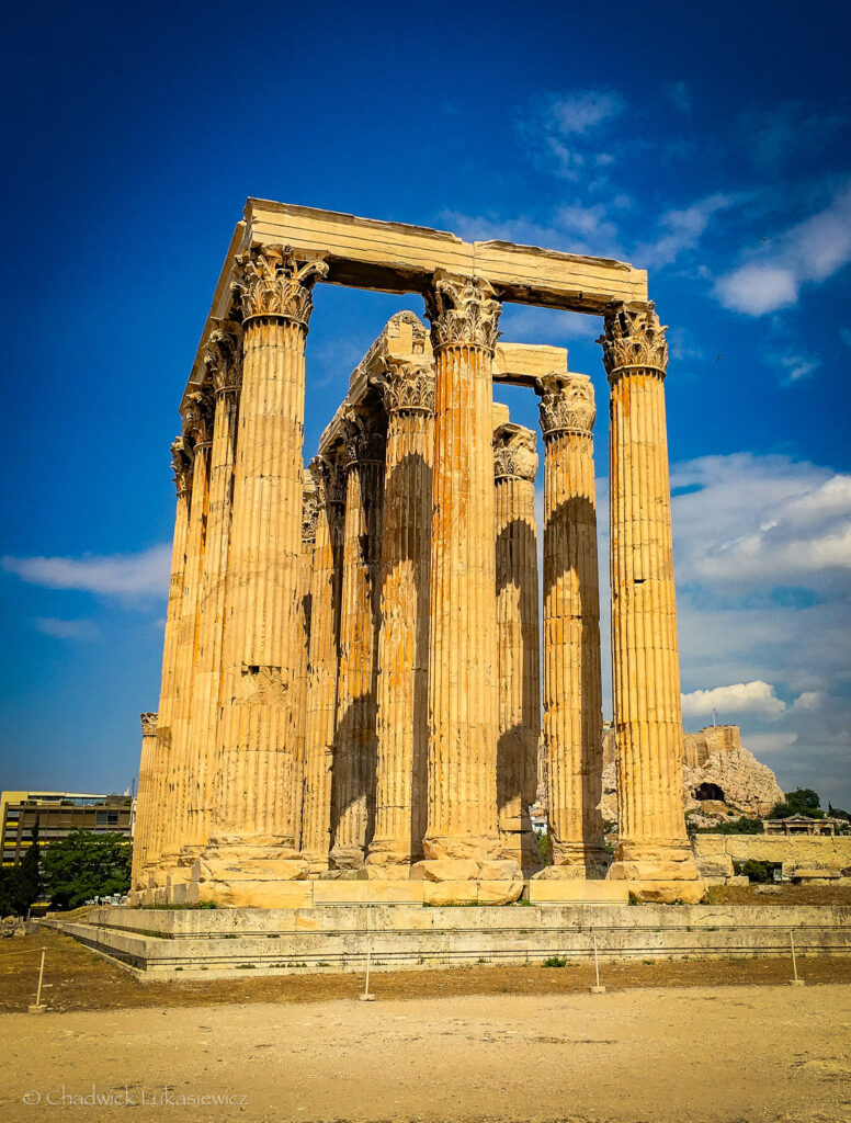 The Temple of Olympian Zeus in Athens, Greece, featuring towering Corinthian columns that rise dramatically against a bright blue sky. The golden tones of the weathered marble are illuminated by sunlight, showcasing the intricate details of the column capitals. In the background, the Acropolis and nearby modern buildings provide a contrast between ancient and contemporary Athens.