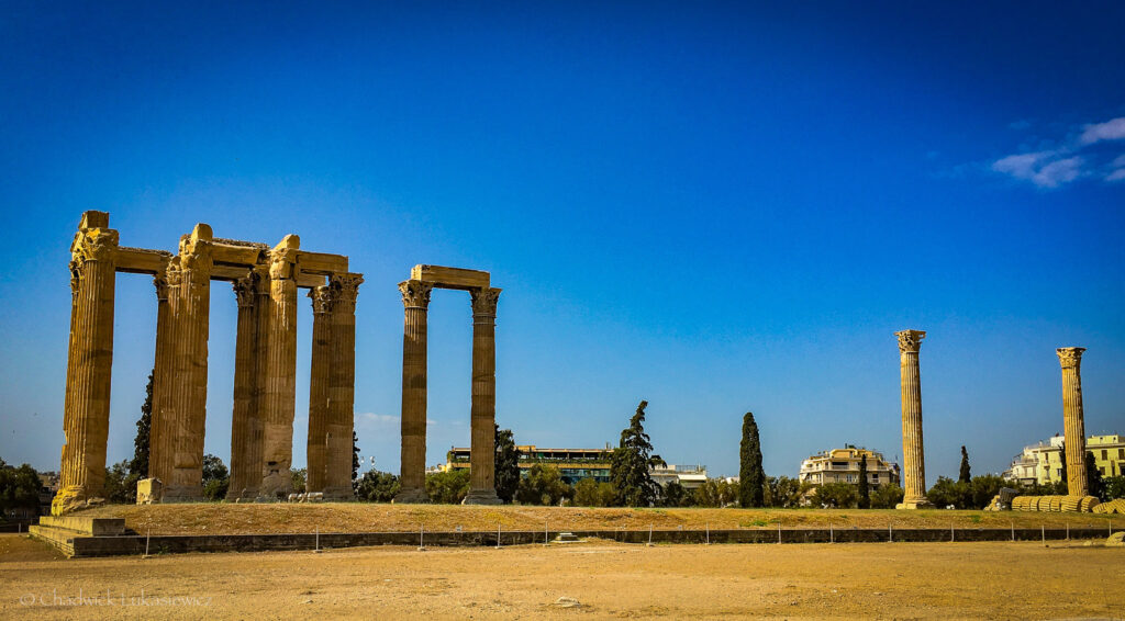 A clear view of the ruins of the Temple of Olympian Zeus in Athens, Greece, set against a bright blue sky. The ancient stone columns, some still standing tall while others lie in ruins, reflect the grandeur of this historic site. Modern buildings and trees in the background highlight the juxtaposition of Athens’ rich history and contemporary city life. A perfect visual for an ADHD travel guide to Rome and Athens, showcasing iconic archaeological marve