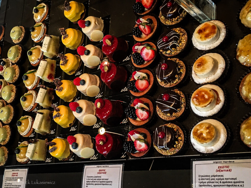 An elegant display of assorted Greek pastries arranged in neat rows at a bakery. The colorful selection includes miniature tarts topped with fresh fruit like strawberries, blackberries, and raspberries, as well as glossy cakes coated with vibrant yellow, red, and white glazes. Some pastries are decorated with chocolate drizzles, nuts, and caramelized sugar. The dark countertop accentuates the bright and enticing colors of the desserts, creating a luxurious and appetizing presentation. Labels with descriptions in Greek are visible at the bottom.