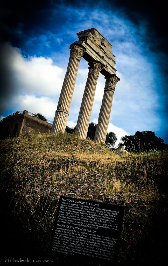 The Temple of Castor and Pollux at the Roman Forum in Rome, Italy, with three tall Corinthian columns supporting a partially preserved architrave. The ruins stand on a grassy slope, surrounded by a sunny sky with scattered clouds. A plaque at the base provides historical information about the temple in Italian and English.