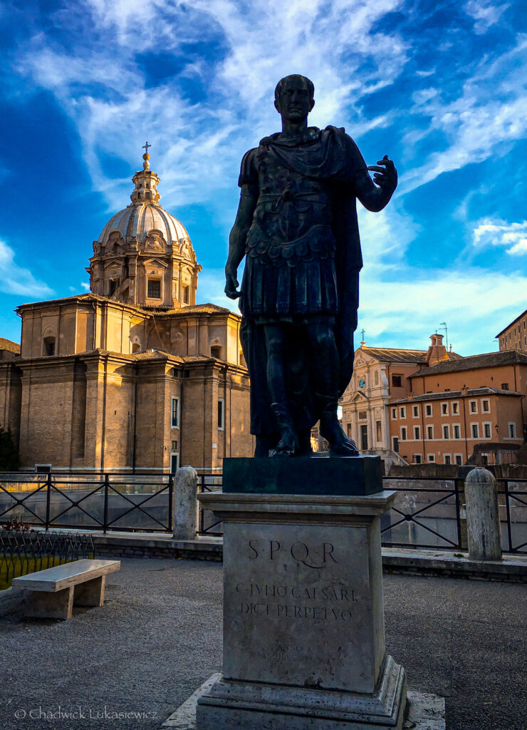 A statue of Julius Caesar standing prominently in front of the Church of Santi Luca e Martina in Rome, Italy. The dark bronze figure of Caesar, dressed in a Roman toga and armor, is silhouetted against the bright dome and warm stone facade of the church. The pedestal below the statue is engraved with the words “S.P.Q.R. C. IVLIO CAESARI DIC PERPETVO.” The vibrant blue sky and scattered clouds enhance the dramatic and historical setting.