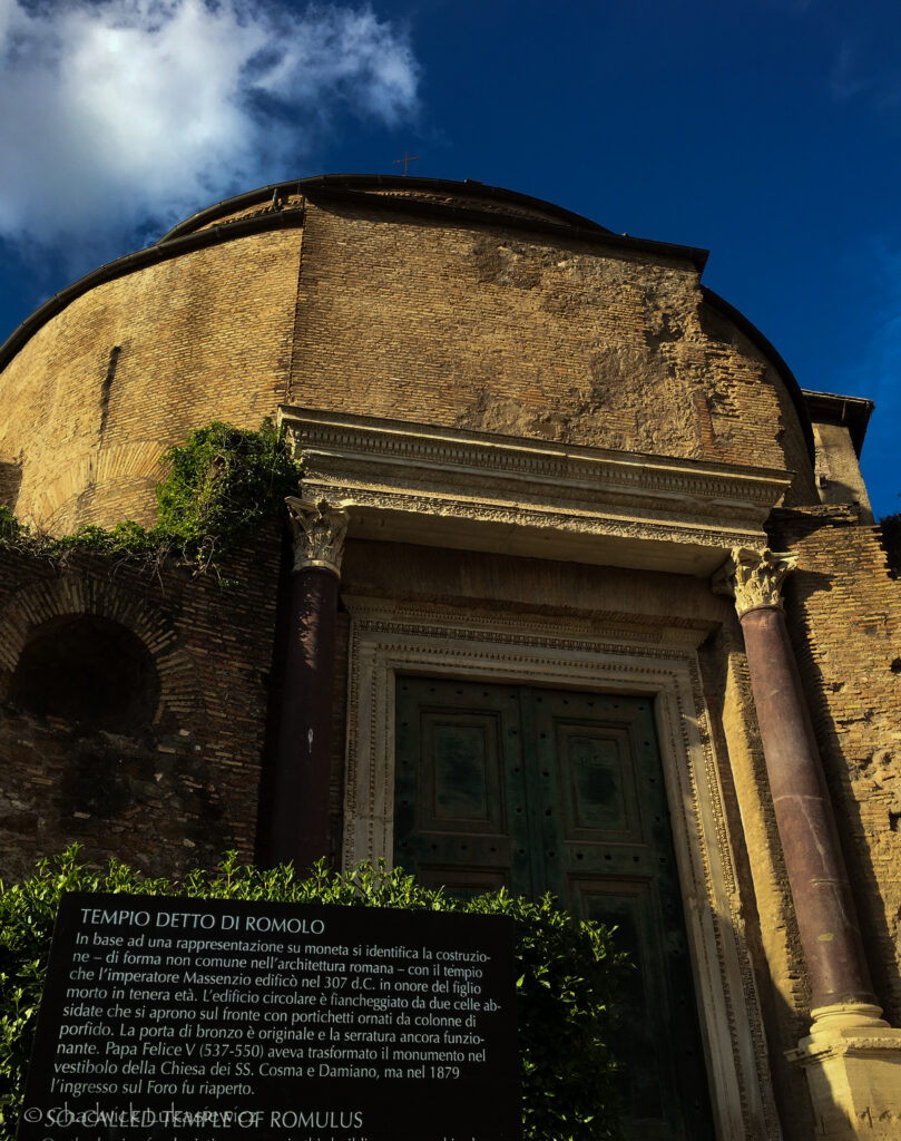 The Temple of Romulus at the Roman Forum in Rome, Italy, featuring a circular brick structure with a prominent green-bronze door framed by intricately carved columns. Ivy grows along parts of the facade, adding a natural touch to the ancient architecture. A clear blue sky and white clouds form a picturesque backdrop, and a plaque in front provides details about the monument.