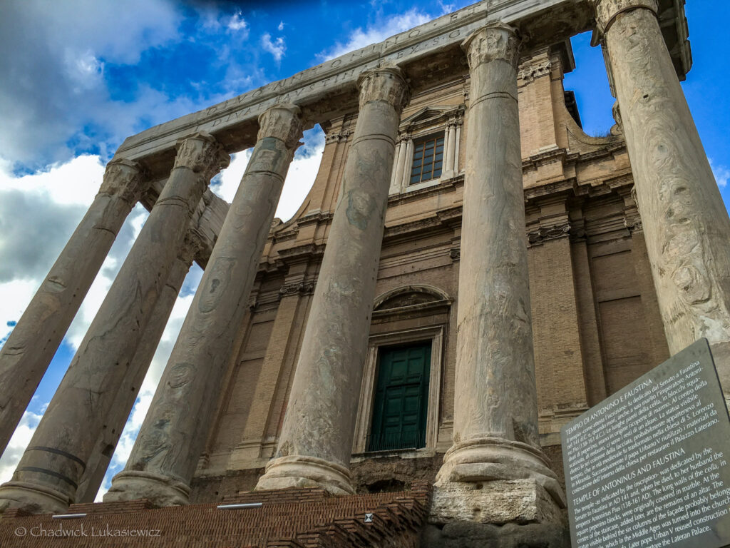 A dramatic upward view of the Temple of Antoninus and Faustina in the Roman Forum, Rome, Italy. The marble columns, adorned with intricate carvings, stand tall against a backdrop of blue sky and clouds, highlighting the grandeur of this ancient Roman structure. An informational plaque in the foreground provides historical context. Perfect for an ADHD travel guide to Rome and Athens, showcasing must-visit ancient landmarks.