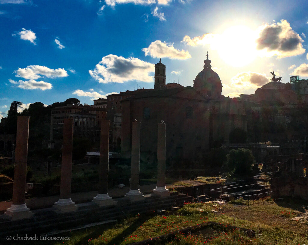 A sunset view of the Roman Forum in Rome, Italy, with the sun casting a golden glow over the historic ruins. In the foreground, a row of ancient columns stands silhouetted against the vibrant sky, while the remnants of stone structures and grassy areas dotted with wildflowers fill the foreground. The skyline includes the dome of a church and other historic buildings, with a prominent statue of a winged horse atop a monument in the distance. The bright sun and scattered clouds create a dramatic and atmospheric contrast, emphasizing the timeless beauty of the scene.