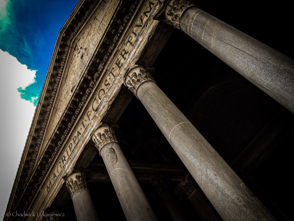 A dramatic, angled view of the Pantheon in Rome, Italy, focusing on its grand portico. The massive Corinthian columns with intricately carved capitals support the triangular pediment, which bears the Latin inscription “M. AGRIPPA L. F. COS TERTIVM FECIT,” commemorating its original builder, Marcus Agrippa. The weathered stone contrasts with the vibrant blue sky and the dark shadowed interior visible in the background, emphasizing the monument’s grandeur and historical significance. The perspective enhances the sense of scale and the timeless elegance of this iconic structure.