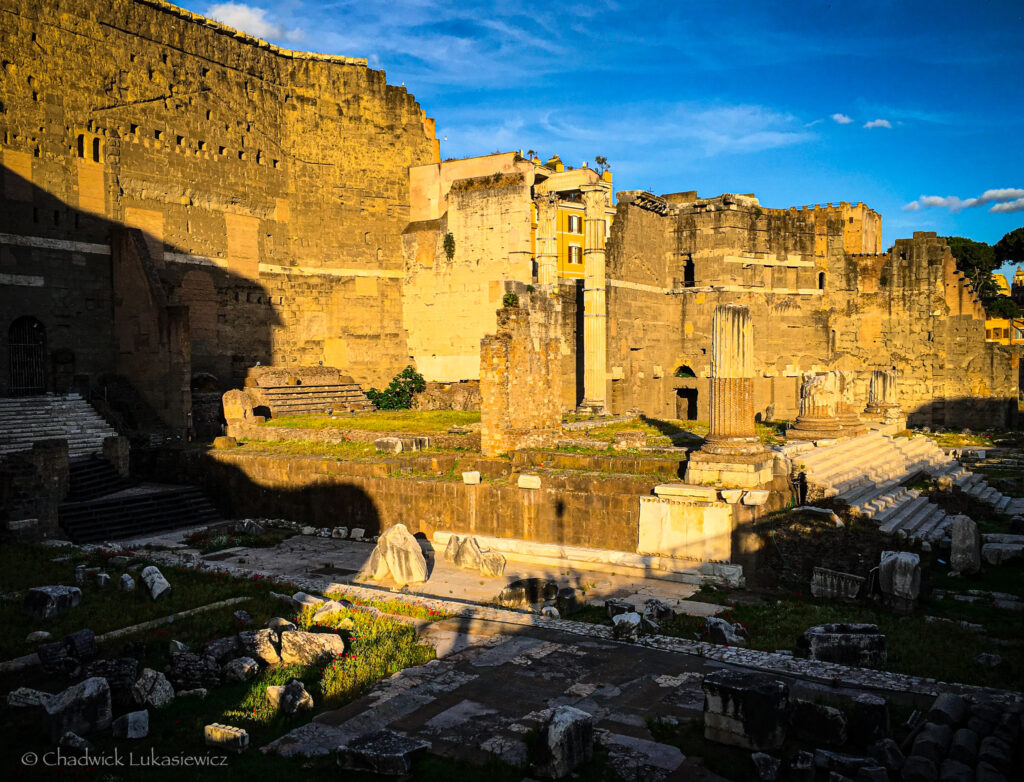 A sunlit view of an ancient Roman ruin, likely part of the Imperial Fora in Rome, Italy. The foreground displays scattered marble fragments and overgrown grass, with a mix of broken columns and remnants of stairs leading to the former structure. The background features towering stone walls with visible erosion and architectural details, alongside partially intact columns and arches. The warm sunlight highlights the textures of the stone, creating a stark contrast with the shaded areas. A modern building peeks out from behind the ruins, blending the ancient and the contemporary.