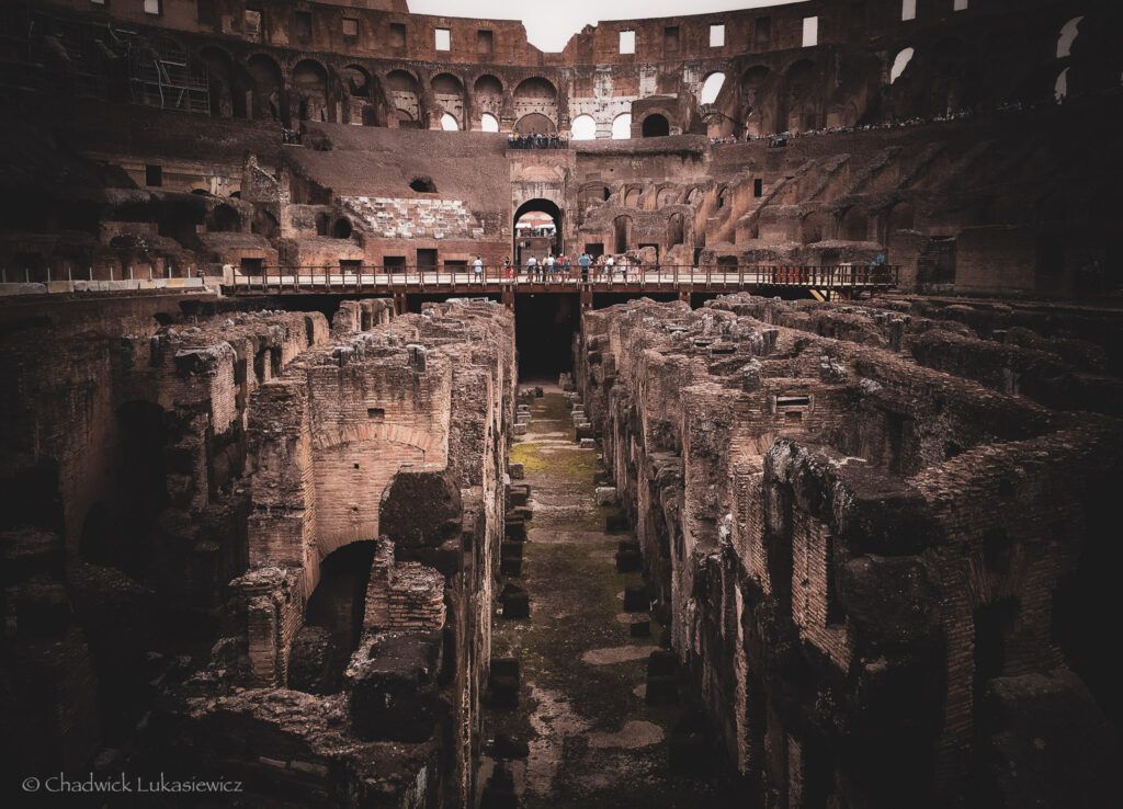 An interior view of the Colosseum in Rome, Italy, showcasing its intricate and weathered substructure. The exposed underground network of walls, corridors, and chambers is visible, where gladiators and animals were once prepared before entering the arena. Above the hypogeum, a modern wooden walkway spans across the ruins, with visitors standing and observing the historical site. The surrounding walls and arches of the amphitheater, though partially eroded, hint at the grandeur of the ancient Roman structure. The soft natural light accentuates the texture and earthy tones of the stone, creating a dramatic and reverent atmosphere.