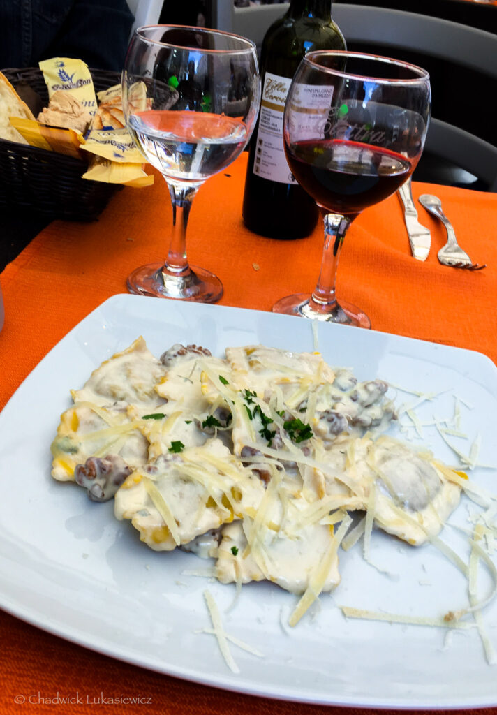 A plate of freshly prepared ravioli served in a creamy white sauce, garnished with shaved parmesan cheese and a sprinkle of parsley. The dish is set on an orange tablecloth with two glasses—one filled with water and the other with red wine—next to a bottle of wine. A bread basket with prepackaged crackers is visible in the background, adding to the rustic Italian dining ambiance.