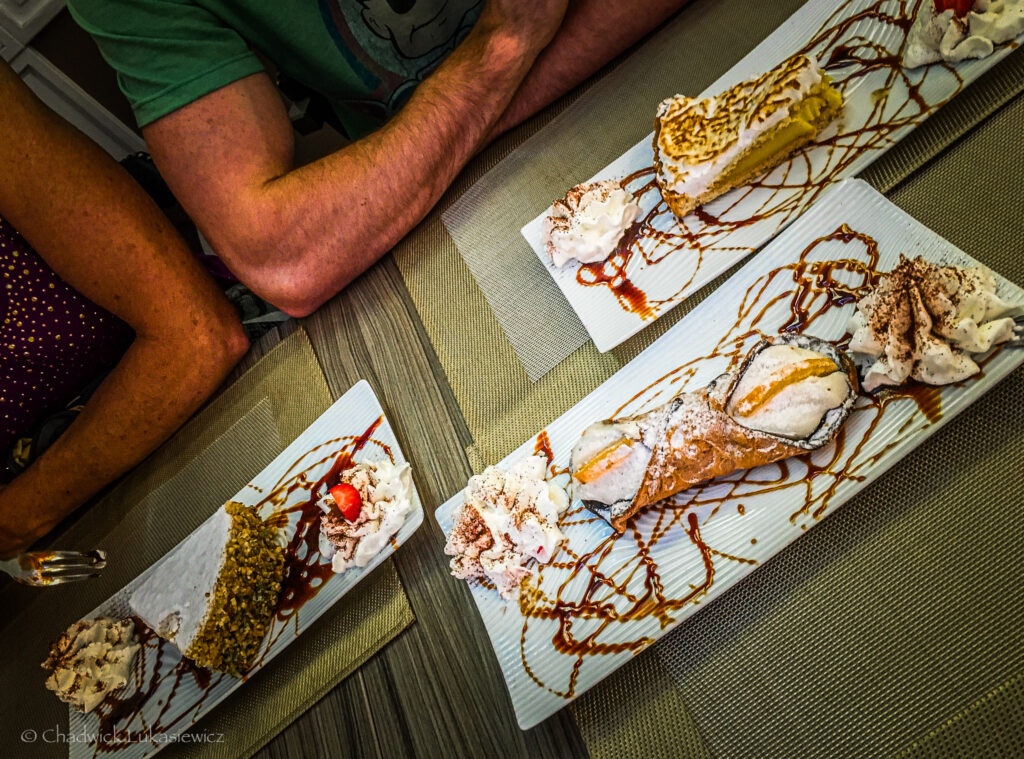 A decadent assortment of Italian desserts elegantly presented on rectangular plates. The foreground features a crisp cannoli dusted with powdered sugar, accompanied by dollops of whipped cream and drizzled caramel sauce. Additional plates hold slices of layered cakes topped with whipped cream and similar caramel drizzle. The warm tones of the wooden table and the casual presence of diners’ arms create a cozy, indulgent dining atmosphere.