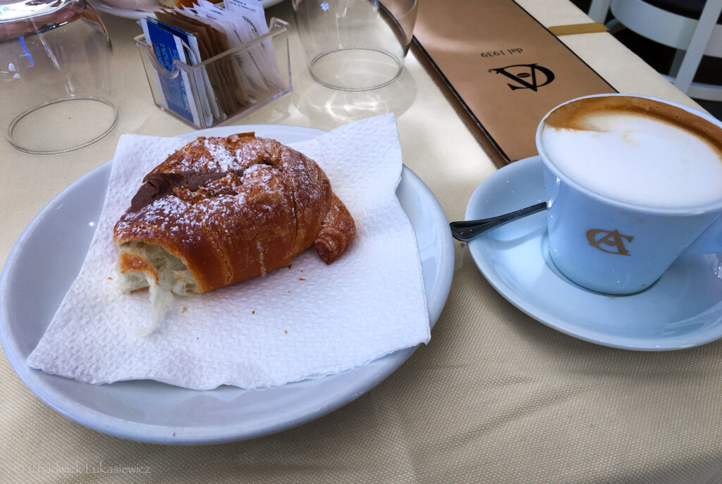 A classic Italian breakfast featuring a partially eaten croissant dusted with powdered sugar, resting on a white napkin on a plate. To the right is a cappuccino with a thick layer of foam served in a branded white cup with a saucer. The table is set with a menu, sugar packets in a glass holder, and clear glasses, creating a simple yet refined morning dining experience.