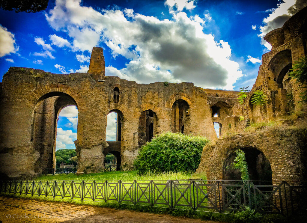 A vibrant view of ancient Roman ruins at Palatine Hill in Rome, Italy. The stone walls and arched openings of the crumbling structure are framed against a bright blue sky with fluffy white clouds. Overgrown vegetation, including green grass and shrubs, adds a touch of nature to the historic site. A black metal fence runs along the foreground, emphasizing the preserved and tranquil setting of this archaeological landmark.