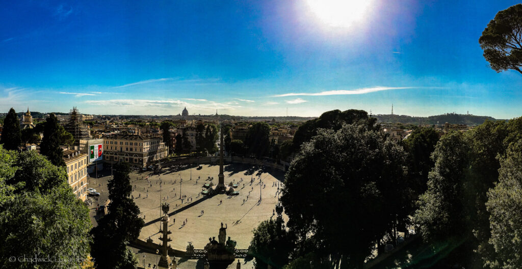 A panoramic view of Piazza del Popolo in Rome, Italy, as seen from the Pincio Terrace. The large oval-shaped square is framed by ornate fountains, statues, and an ancient Egyptian obelisk at its center. People are scattered across the piazza, casting long shadows under the bright afternoon sun. The surrounding cityscape stretches out into the distance, with domes, rooftops, and greenery blending harmoniously. In the background, St. Peter’s Basilica is visible on the horizon, with the clear blue sky above creating a vibrant and expansive atmosphere.