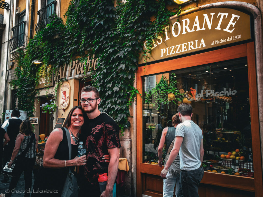 A cheerful street view of “Ristorante Pizzeria al Picchio” in Rome, Italy. A smiling woman and a man pose together in front of the restaurant, which features a golden-yellow exterior covered with lush green ivy. The restaurant’s large windows reflect the street scene and display fresh fruits inside. Other pedestrians are visible along the narrow street, contributing to the lively and cozy atmosphere of the Roman neighborhood.