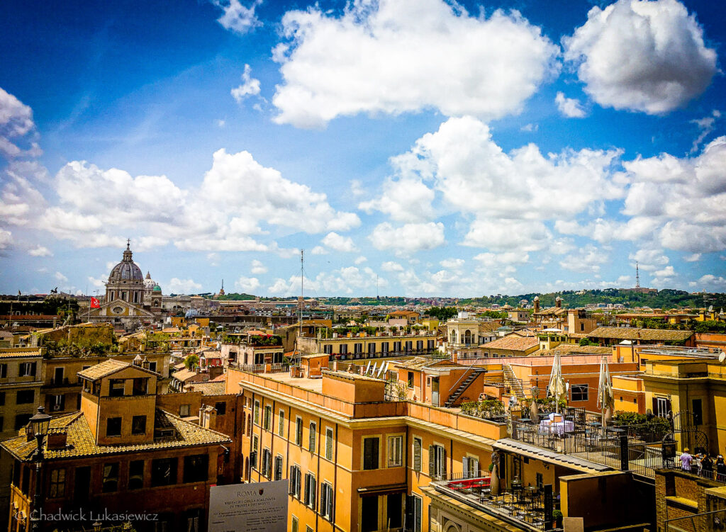 A panoramic rooftop view of Rome, Italy, showcasing the city’s distinctive terracotta and pastel-toned buildings under a bright blue sky filled with fluffy white clouds. The dome of St. Peter’s Basilica rises prominently on the left, adding a historic landmark to the vibrant cityscape. Rooftop terraces adorned with umbrellas and greenery dot the foreground, while rolling green hills stretch into the distance, blending the urban and natural beauty of Rome.