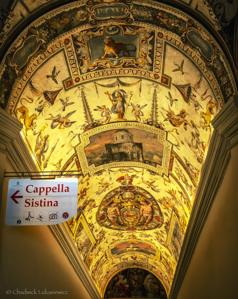 An ornate, brightly illuminated arched ceiling leading to the Sistine Chapel in Vatican City. The ceiling is richly decorated with detailed frescoes depicting biblical and mythological figures, cherubs, and intricate floral patterns, framed with golden accents. A prominent scene in the center features a classical building surrounded by angels, with additional vibrant medallions and motifs along the arch. On the left, a directional sign reads “Cappella Sistina” with icons indicating accessibility and photography restrictions. The scene captures the grandeur and artistry of Renaissance design, guiding visitors towards one of the world’s most famous religious landmarks.