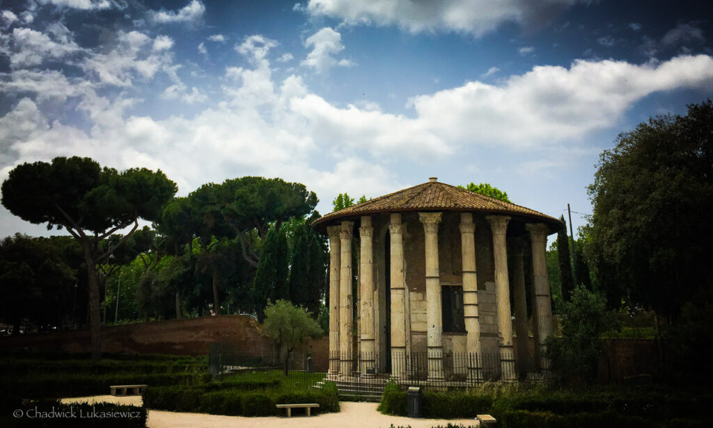 A serene daytime view of the Temple of Hercules Victor in Rome, Italy. The ancient circular temple is surrounded by tall Corinthian marble columns and capped with a terracotta-tiled conical roof. It stands within a landscaped park, framed by lush greenery, including neatly trimmed hedges, tall pine trees, and cypress trees. The bright blue sky with soft white clouds adds a tranquil backdrop to this historical setting. A small stone bench is visible in the foreground, enhancing the sense of quiet and reflection in this iconic Roman site.