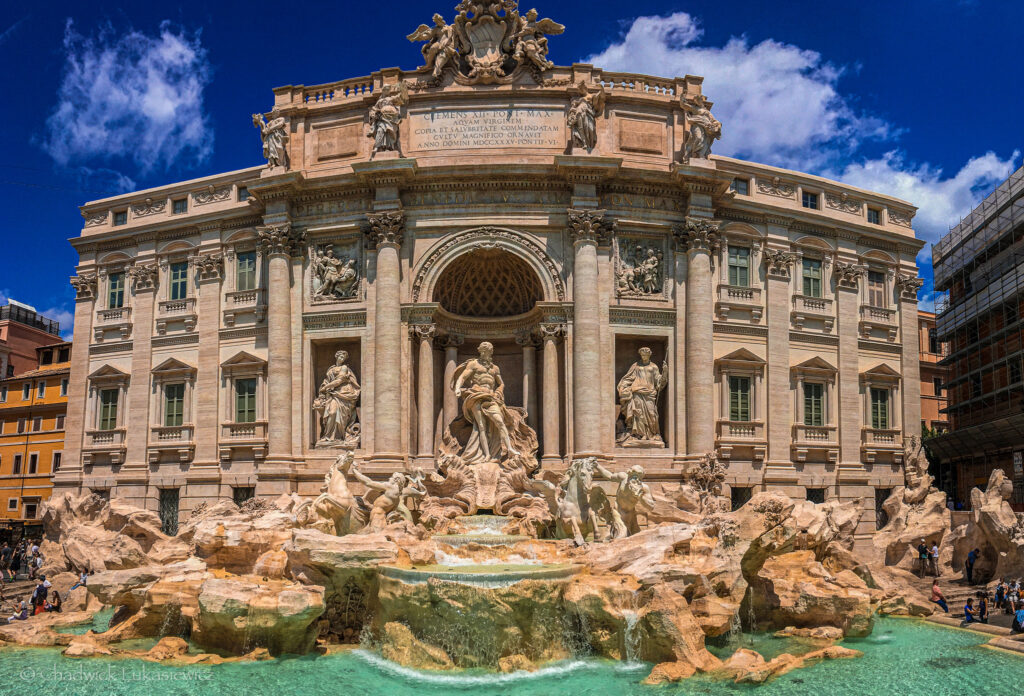 A stunning view of the Trevi Fountain in Rome, Italy, a monumental baroque masterpiece. The central figure is Oceanus, standing atop a chariot drawn by sea horses, surrounded by allegorical statues and cascading waterfalls. The intricately detailed sculptures emerge from rugged stone, creating the illusion of a natural grotto. The surrounding facade is adorned with Corinthian columns, niches containing statues, and an ornate inscription at the top. The turquoise water in the basin reflects the bright sunlight under a vibrant blue sky with scattered white clouds. Tourists are gathered around the edges, admiring and photographing the iconic landmark.