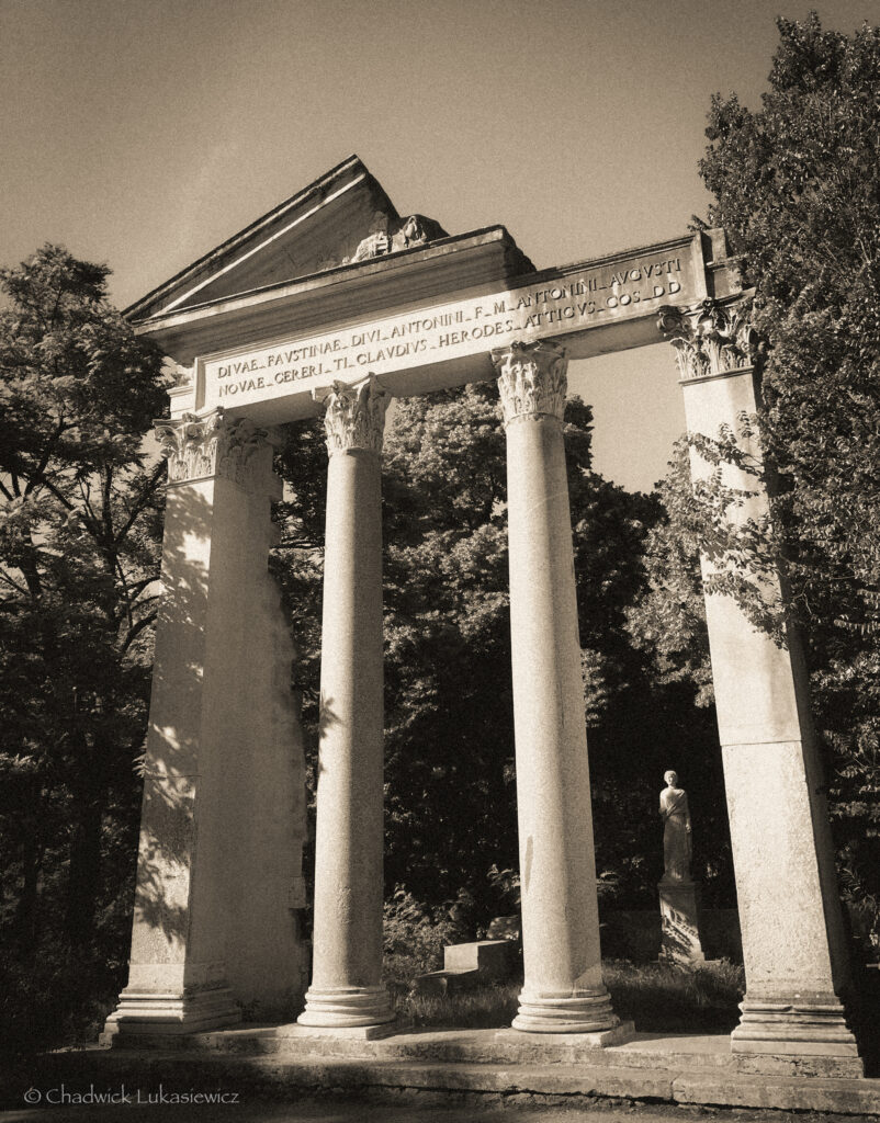 A sepia-toned photograph of a neoclassical temple in the Gardens of the Villa Borghese, Rome, Italy. Four tall columns with ornate capitals support a triangular pediment inscribed with text. Behind the columns, a statue of a robed figure is visible, surrounded by lush trees. The sepia effect gives the image a vintage and timeless quality.