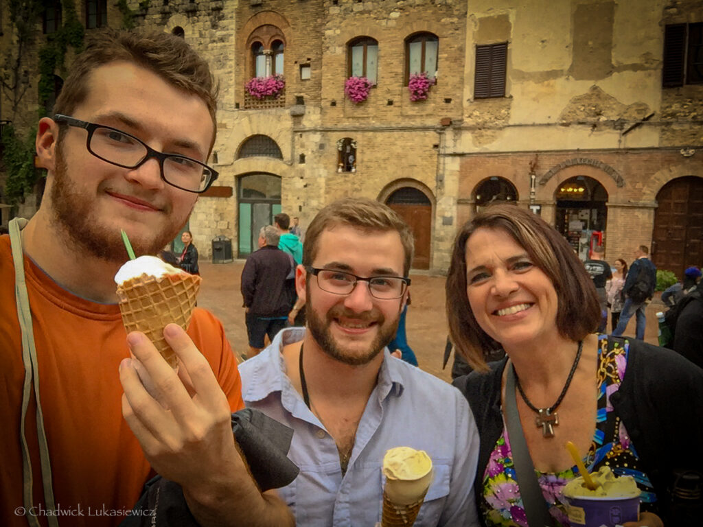 A joyful group photo featuring three people holding gelato in the medieval town of San Gimignano, Italy. The man on the left holds a waffle cone with a scoop of gelato topped with a small green spoon, while the man in the center and the woman on the right also enjoy their gelato, smiling warmly. Behind them, the backdrop showcases historic stone buildings adorned with vibrant pink flowers in window boxes, contributing to the charming, old-world atmosphere of the square. Other visitors can be seen exploring the picturesque surroundings.