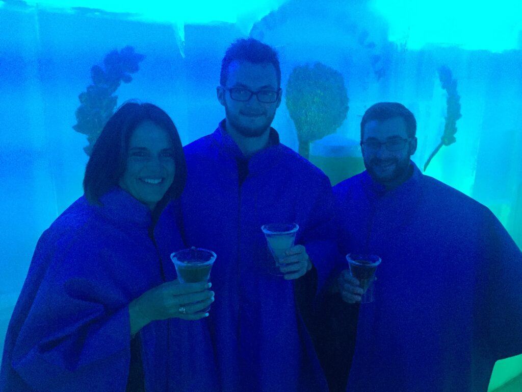 Three individuals standing close together in a room with a bright blue hue, likely an ice bar. They are wearing matching blue cloaks, holding cocktails in transparent glasses. The icy walls and frozen decor in the background suggest a themed environment. The group is smiling and appears to be enjoying the unique, chilled atmosphere.