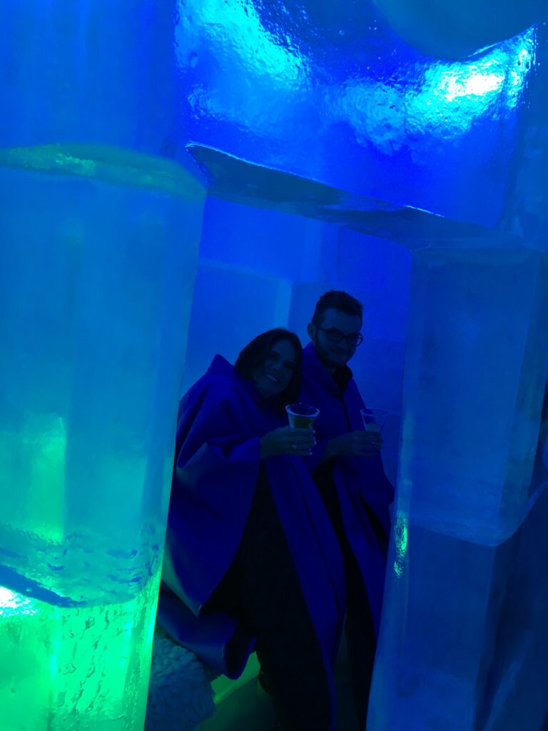 Two people, a woman and a man, seated inside a vibrant blue-lit room with icy structures around them, likely an ice bar. They are dressed in blue cloaks and holding drinks in clear glasses. A large block of illuminated ice with a green glow is visible in the foreground, adding to the frosty ambiance. The setting has an immersive and futuristic vibe due to the lighting and reflective surfaces.