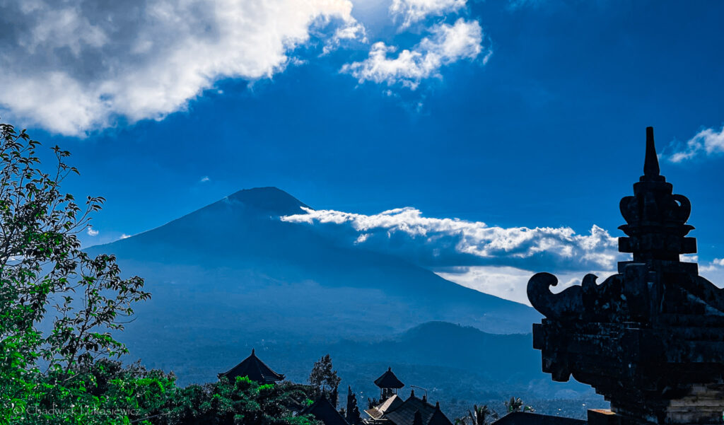 A majestic view of Mount Agung, Bali’s sacred volcano, partially veiled in misty clouds, framed by ornate temple rooftops and lush greenery under a deep blue sky.