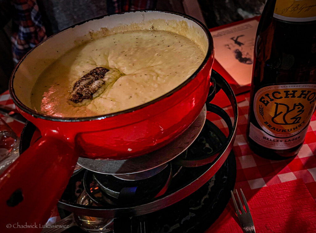 A close-up of a red pot filled with creamy, melted cheese fondue on a tabletop burner, accompanied by a bottle of Eichhof Helles Lager beer on a checkered red-and-white tablecloth.