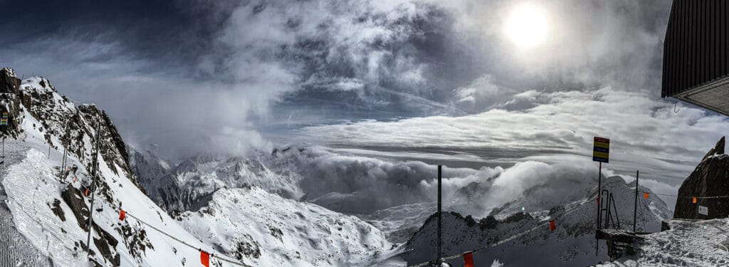 A panoramic view from the top of Gemsstock at Andermatt-Sedrun-Disentis in Switzerland, showcasing rugged snow-covered peaks under a dramatic, partly cloudy sky. Snowboarding trails are marked with orange flags and safety signs, providing a visual setting for ADHD snowboarding tips to navigate challenging alpine terrain while maintaining focus and safety.
