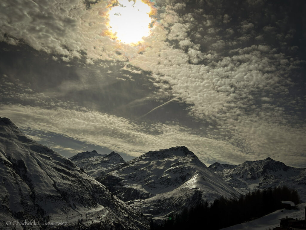 A moody alpine sunset in Andermatt, Switzerland, with snow-covered peaks silhouetted under a cloudy sky, as the sun peeks through with a golden glow.