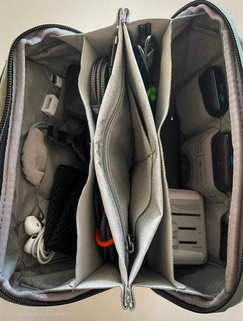 An overhead view of an open Peak Design Tech Pouch in sage green, showcasing its neatly organized interior compartments. The pouch contains an array of tech accessories, including earbuds, cables, adapters, and a white universal travel adapter. The structured gray interior emphasizes efficient storage, with separate slots and mesh sections for easy access to items.