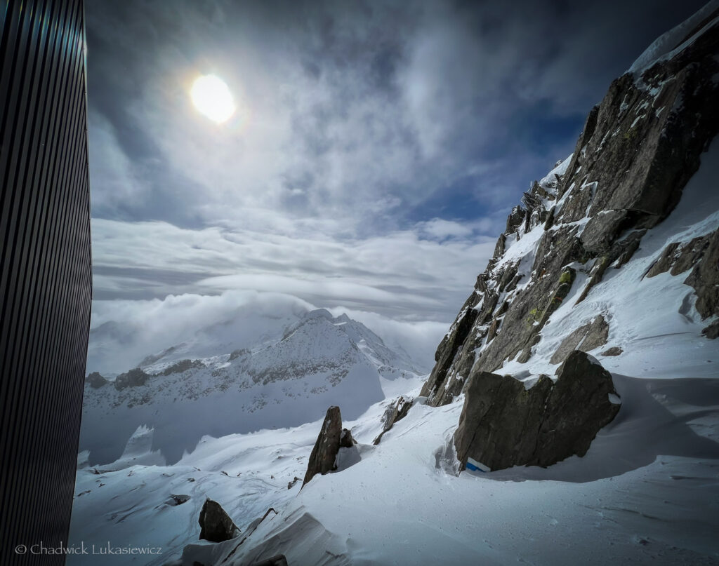 A breathtaking view from Gemsstock in Andermatt, Switzerland, with jagged rocky cliffs and snow-covered slopes leading into a misty alpine valley under a hazy sun.