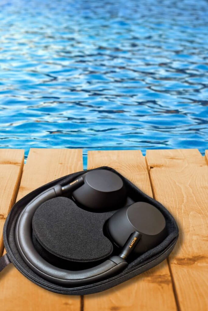 Black Sony WH-1000XM5 headphones neatly folded inside their carrying case, resting on a wooden deck next to a vibrant blue pool