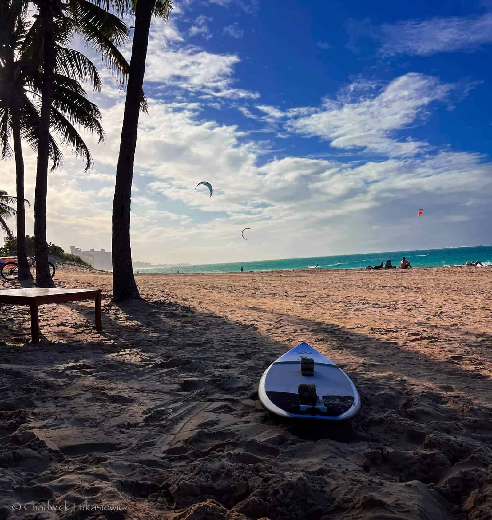 A surfboard resting on the sandy beach under palm trees in Puerto Rico, with kite surfers in the distance and a bright blue sky filled with clouds.