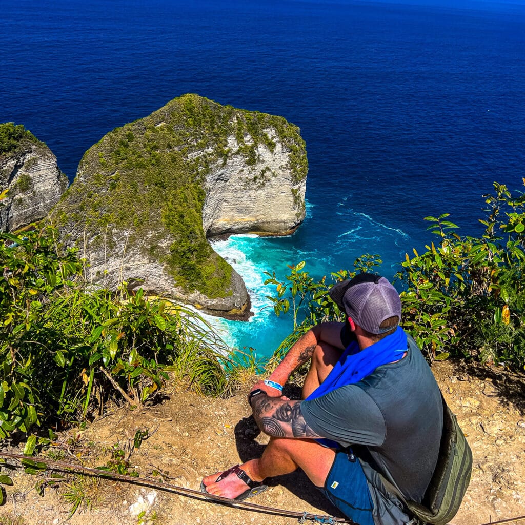 A travel photographer sits at Kelingking Beach overlook in Bali, showcasing the importance of ADHD-friendly travel photography gear for neurodivergent creatives