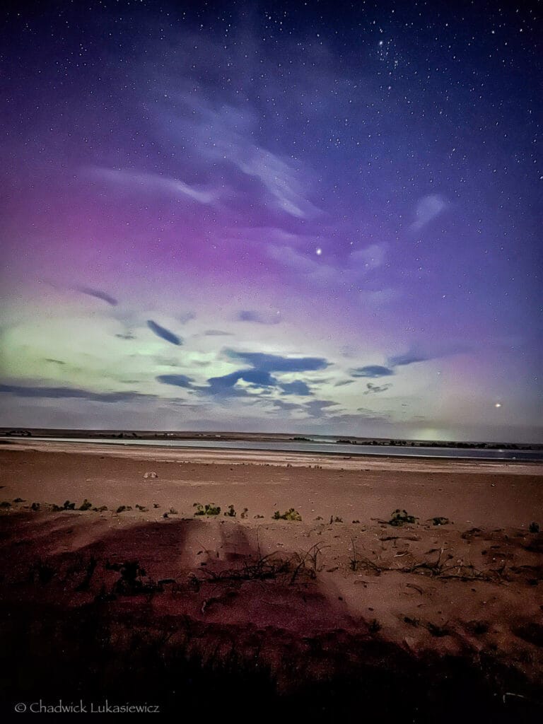 Purple and green aurora borealis over Jackson Lake, Colorado, with a star-filled sky and barren landscape below.