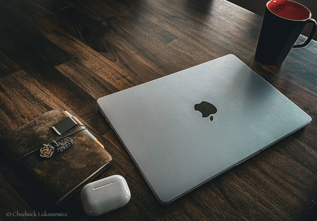 A MacBook, Traveler’s Notebook, wireless earbuds, and a coffee mug on a wooden desk, showcasing a simple ADHD-friendly editing setup for travel creatives.
