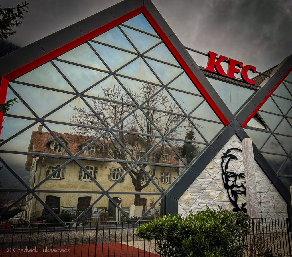 A distorted reflection of a historic Swiss building in the modern glass facade of a KFC in Chur, Switzerland, illustrating the contrast between perfectionism and creative imperfection.