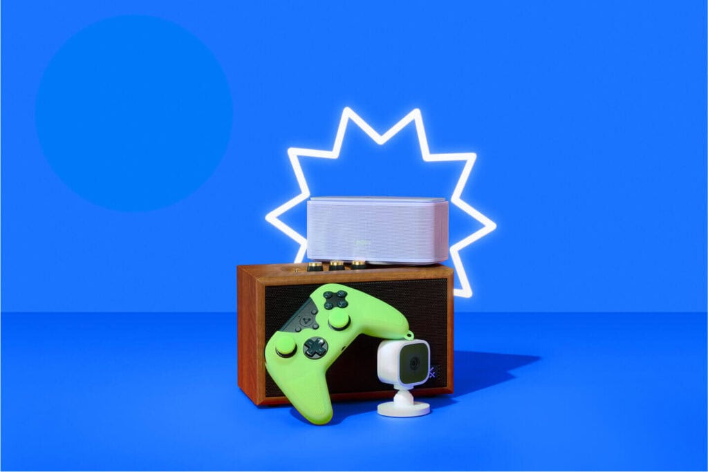 A flat-lay of Prime Day tech deals against a vibrant blue background with a glowing white starburst outline: a wooden retro speaker topped by a sleek white Bluetooth speaker, laid in front of a neon-green wireless game controller and a compact white cube action camera on a stand