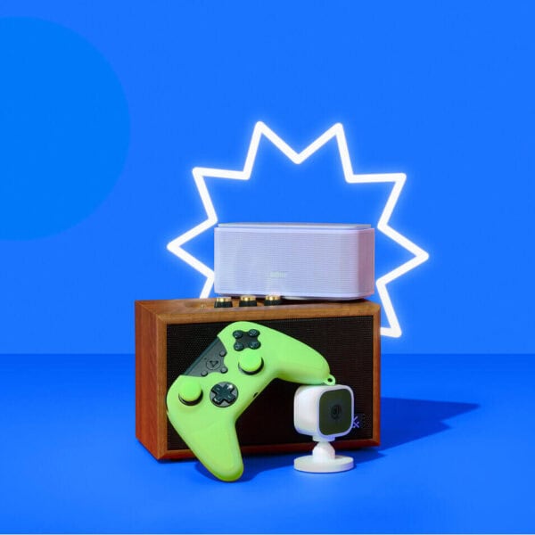 A flat-lay of Prime Day tech deals against a vibrant blue background with a glowing white starburst outline: a wooden retro speaker topped by a sleek white Bluetooth speaker, laid in front of a neon-green wireless game controller and a compact white cube action camera on a stand