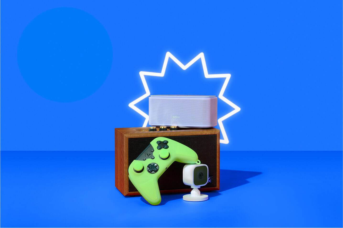 A flat-lay of Prime Day tech deals against a vibrant blue background with a glowing white starburst outline: a wooden retro speaker topped by a sleek white Bluetooth speaker, laid in front of a neon-green wireless game controller and a compact white cube action camera on a stand