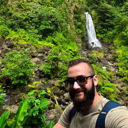 a man with a beard and sunglasses on a mountain by a waterfall